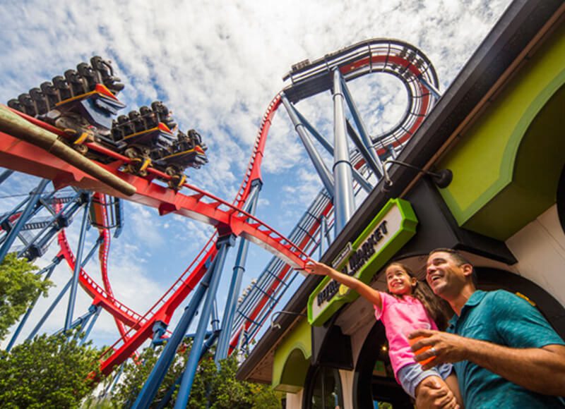 A low-angle shot of a father carrying his daughter under roller coaster tracks.